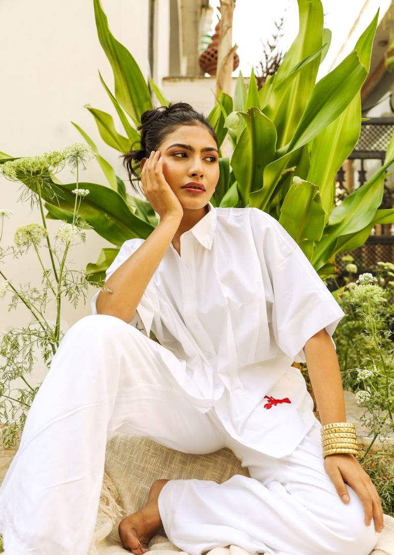 A person wearing an oversized white shirt with wide sleeves and a red lobster patch on the hem, sitting in a relaxed pose with one hand touching their face, surrounded by green plants.