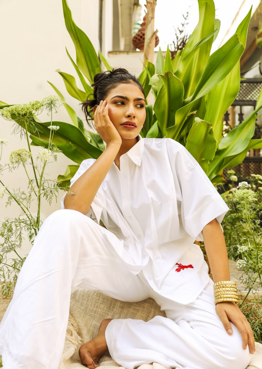 A person wearing an oversized white shirt with wide sleeves and a red lobster patch on the hem, sitting in a relaxed pose with one hand touching their face, surrounded by green plants.